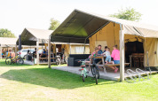 Familie genießt Zeit vor einem Safarizelt im Kampeerdorp de Zandstuve, Niederlande, mit Fahrrädern und Tischen.