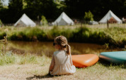 Lille pige sidder på græsset nær REBL Outdoor campingplads i Friesland med telte i baggrunden.