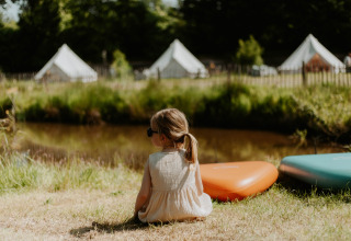 Lille pige sidder på græsset nær REBL Outdoor campingplads i Friesland med telte i baggrunden.