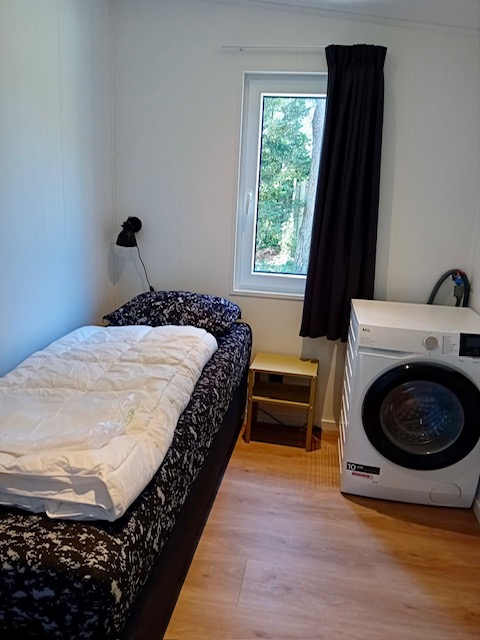 Piccola camera alla Forest Lodge De Mooie Plek con letto singolo, lavatrice e finestra che guarda il bosco.