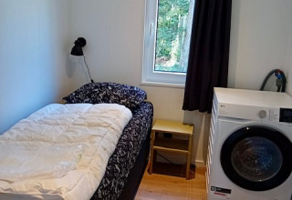 Piccola camera alla Forest Lodge De Mooie Plek con letto singolo, lavatrice e finestra che guarda il bosco.