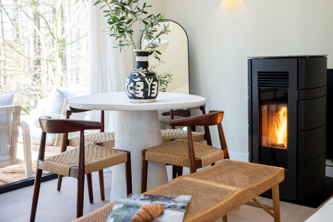 Cozy dining area with woven chairs, round table, vase and fireplace in Koya Tiny Cabin, Wilsumer Berge, Germany.