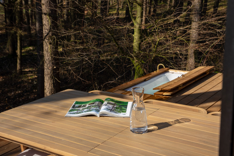 Table en bois extérieure avec carafe d'eau et magazine, vue sur forêt et baignoire à Koya Tiny Cabin, Wilsumer Berge.