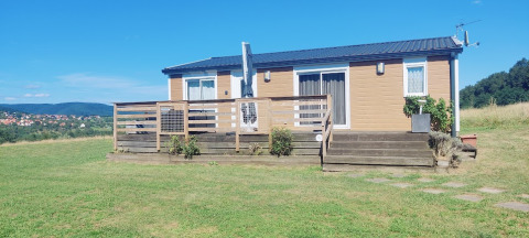Un lodge de una planta con terraza de madera, rodeado de campos verdes y cielo azul despejado.