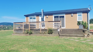 Un lodge de una planta con terraza de madera, rodeado de campos verdes y cielo azul despejado.