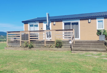 Un lodge de una planta con terraza de madera, rodeado de campos verdes y cielo azul despejado.