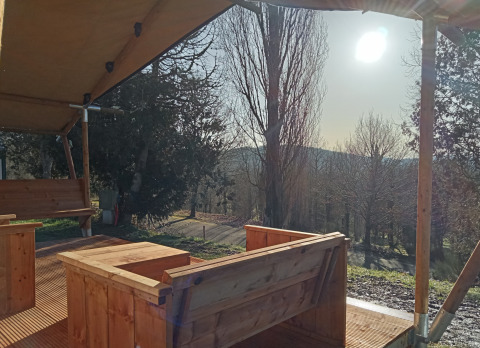 Vista desde una tienda safari en Camping La Perle, Francia, con muebles de madera y luz solar entre árboles.