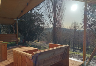 Vista desde una tienda safari en Camping La Perle, Francia, con muebles de madera y luz solar entre árboles.