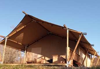 Tienda de safari con estructura de madera y techo de lona situada en la naturaleza bajo cielo azul claro.