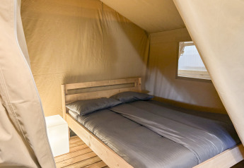 Vista interna di una tenda safari al Camping La Perle in Francia con letto matrimoniale e comodino bianco.