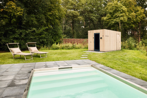 Outdoor swimming pool with lounge chairs on a grassy lawn next to a modern wooden lodge surrounded by trees.