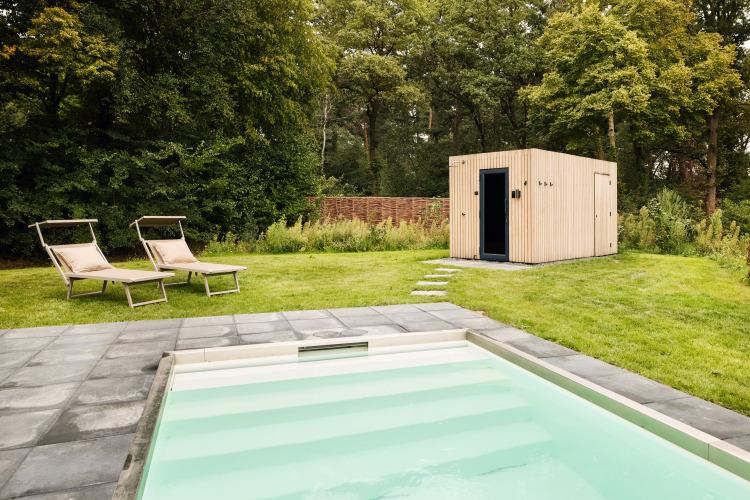 Piscina exterior con tumbonas en el césped junto a una moderna caseta de madera rodeada de árboles y vegetación.