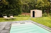 Outdoor swimming pool with lounge chairs on a grassy lawn next to a modern wooden lodge surrounded by trees.