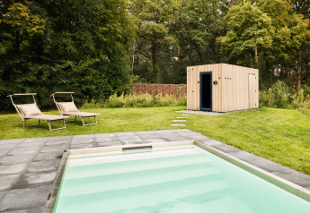 Piscina exterior con tumbonas en el césped junto a una moderna caseta de madera rodeada de árboles y vegetación.