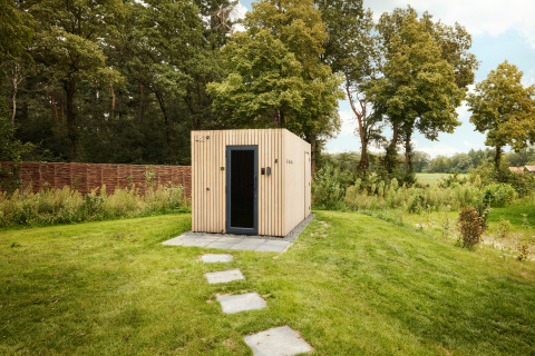 Eine moderne Holzhütte mit schwarzer Tür steht auf einer grünen Wiese, umgeben von Bäumen und Natur.