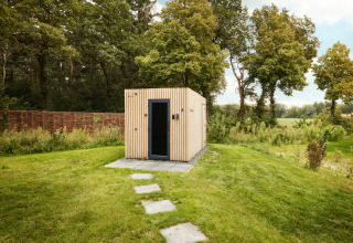 Een modern houten lodge met zwarte deur staat op een grasveld, omringd door bomen en groen.
