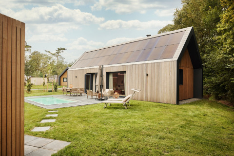 Foto af moderne lodge med store solpaneler på taget, en swimmingpool og møbleret terrasse i grønne omgivelser.