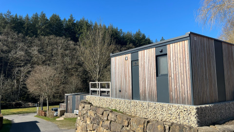 Pequeña casa de madera sobre base de piedra rodeada de bosque y cielo azul en Kohnenhof, Luxemburgo.