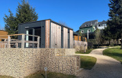 Moderne tiny house ved Kohnenhof i Luxembourg, omgivet af træer, med blå himmel og gangsti foran.