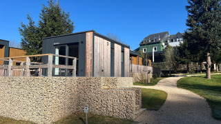 Moderne tiny house ved Kohnenhof i Luxembourg, omgivet af træer, med blå himmel og gangsti foran.