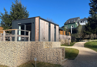 Moderne tiny house ved Kohnenhof i Luxembourg, omgivet af træer, med blå himmel og gangsti foran.
