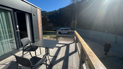 Terraza de madera en Tiny House en Kohnenhof, Luxemburgo, con mesa, sillas, coche y sol brillante al fondo.