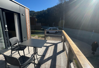 Træterrasse ved Tiny House i Kohnenhof, Luxembourg, med bord, stole, bil og solskin i baggrunden.