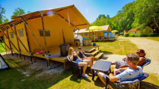 Familie nyder solskin foran en Lodgetent Bordeaux safaritelt på en campingplads omgivet af natur.