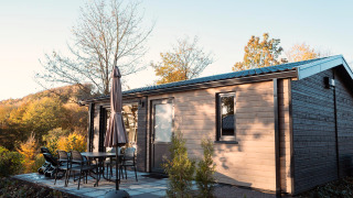 Outdoor photo of a modern wooden lodge with patio furniture, umbrella, and trees in a scenic landscape.