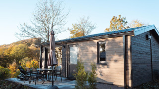 Outdoor photo of a modern wooden lodge with patio furniture, umbrella, and trees in a scenic landscape.
