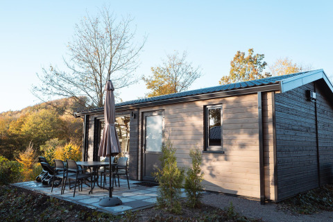 Buitenopname van een moderne houten lodge met terrasmeubilair en parasol, omgeven door natuur.