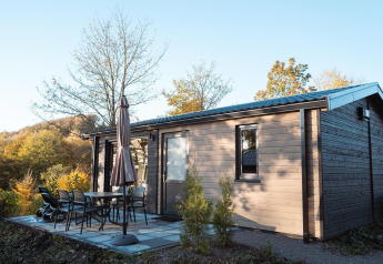 Buitenfoto van een moderne houten lodge met terras, tafel, stoelen, parasol en omringende bomen.