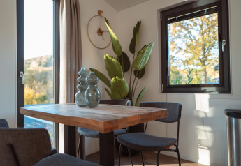 Licht eetgedeelte in een lodge met houten tafel, moderne stoelen, grote ramen en hoge groene plant.