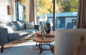 Hyggelig lodge-stue med store vinduer, grå sofa, sofaborde og udsigt til terrasse og natur.