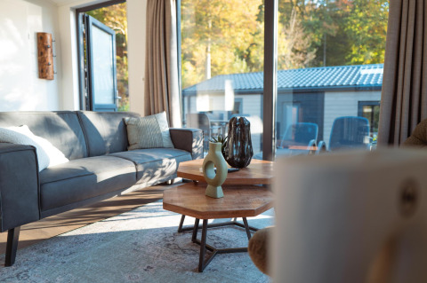 Gemütliches Wohnzimmer in einer Lodge mit grauem Sofa, Glastüren und Blick nach draußen auf Bäume.