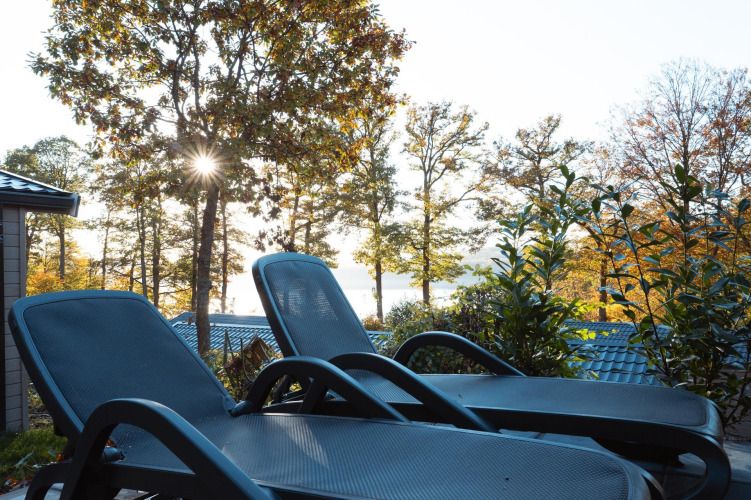 Two lounge chairs on a patio overlooking trees and a lake, with sunlight streaming through the leaves.