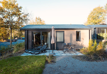 Moderne lodge med terrasse, liggestole, bord og stole i grønne omgivelser under efterårstræer.