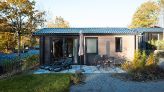 Moderna cabaña con terraza, tumbonas y mesa con sillas, rodeada de árboles en un entorno otoñal.