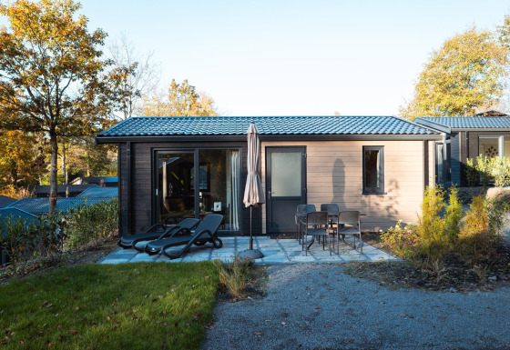 Moderna cabaña con terraza, tumbonas y mesa con sillas, rodeada de árboles en un entorno otoñal.