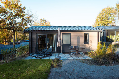 Moderna cabaña con terraza, tumbonas y mesa con sillas, rodeada de árboles en un entorno otoñal.