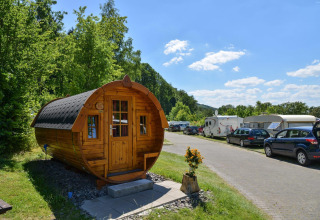 Cabaña tractor en Biggesee, Alemania, rodeada de caravanas, autos y naturaleza bajo un cielo despejado.