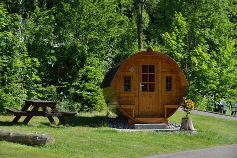 Cabaña de madera con forma de barril en Biggesee, Alemania, rodeada de vegetación y mesa de picnic.
