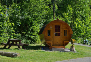 Cabaña de madera con forma de barril en Biggesee, Alemania, rodeada de vegetación y mesa de picnic.