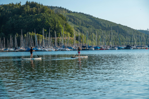 Dos personas haciendo paddle surf en el lago, con muchos veleros y colinas boscosas en Biggesee, Alemania.