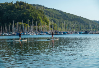Dos personas haciendo paddle surf en el lago, con muchos veleros y colinas boscosas en Biggesee, Alemania.