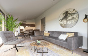 Sala moderna en una cabaña con sofá gris, planta grande, arte de pared redondo y cocina al fondo.