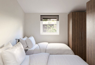 Een lichte lodgekamer met twee eenpersoonsbedden, wit beddengoed, venster en houten kleerkast.