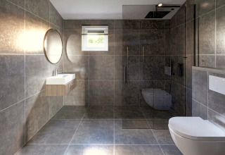 Moderne badkamer in een lodge met grijze tegels, wandtoilet, inloopdouche en een ronde spiegel boven de wastafel.