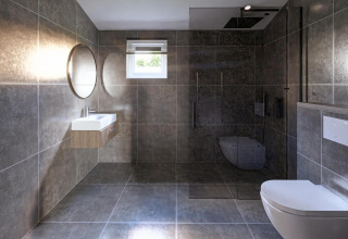 Baño moderno en una cabaña con azulejos grises, inodoro suspendido, ducha abierta y espejo redondo.