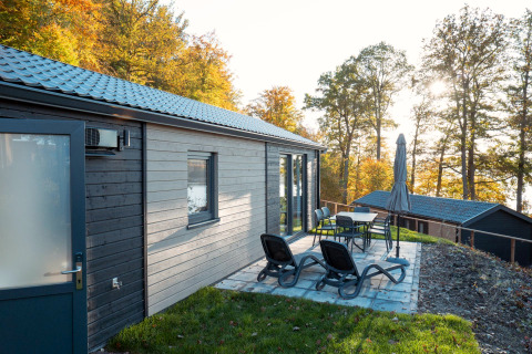 Patio with lounge chairs and dining set outside Hackfort lodge, surrounded by autumnal forest trees.